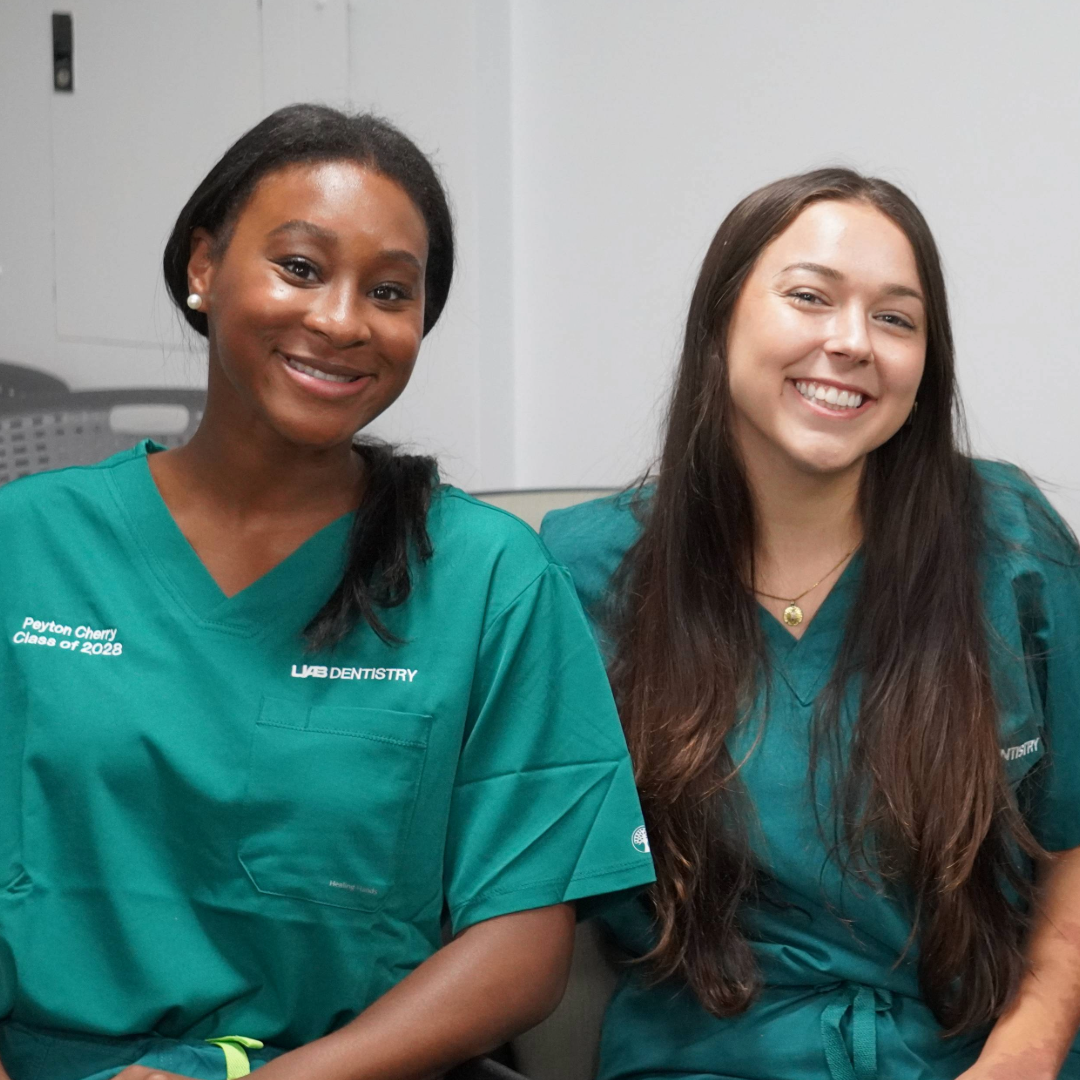 Two girls in green scrubs sitting in a clinical setting smiling for a photo